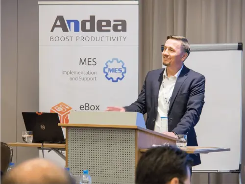 Man in a dark suit smiles while presenting Andea's MES implementation and support solutions at a manufacturing industry event. A laptop and glass of water are on the podium.