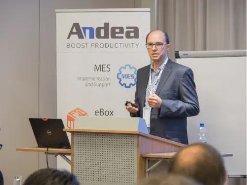 A man in a suit and glasses presents at a podium with a laptop and an Andea banner promoting MES solutions. Attendees are visible in the foreground.