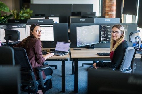 Two women work at computer workstations in a modern office. One focuses on a laptop and spreadsheet on her monitor, the other reviews code on dual monitors. Both look towards the camera.