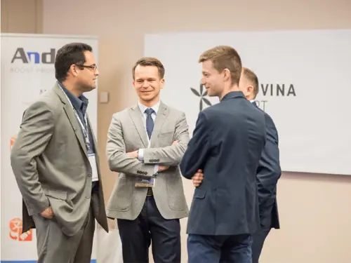 Three men in suits and blazers stand together, engaged in conversation at a business event. One man to the left has his hand in his pocket, while the other two have their arms crossed.
