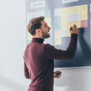 A man in a maroon turtleneck writes on a sticky note on a Kanban board, organizing tasks for agile manufacturing execution systems (MES) or advanced planning and scheduling (APS).