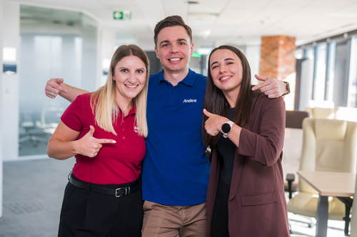 Three smiling Andea team members, two women and one man, posing together in a modern office. They are all pointing at the camera. The man wears a blue polo shirt with the Andea logo.