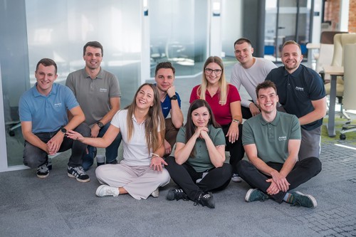 A diverse team of nine smiling people, posing on a carpeted floor in an office setting, wear matching Anda branded polo shirts, showcasing their collaborative spirit in digital manufacturing.