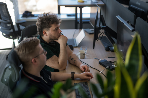 Two colleagues focus on computer screens in a bright office, discussing production data. One man with curly hair rests his chin in his hand, while the other looks intently at a monitor displaying software possibly used for MES or APS.