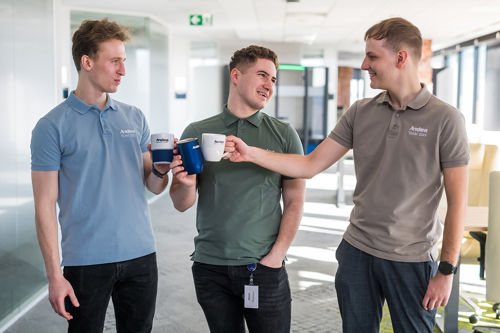 Three smiling men in light blue, green, and grey Andea Team 2023 polo shirts toast with white and blue mugs in a modern office setting.