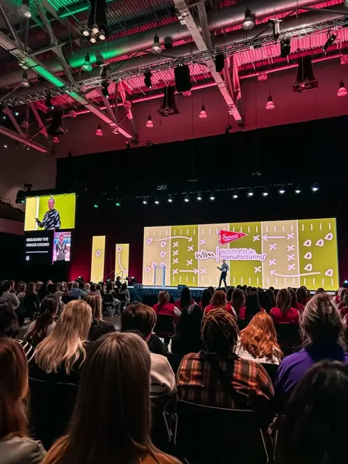 Attendees watch a speaker on stage at the Women in Manufacturing summit. A large screen displays a football play diagram and the summit logo. Green and red stage lighting illuminates the venue.