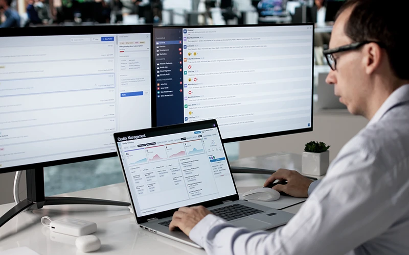 A man in a white shirt and glasses uses a laptop displaying a manufacturing quality management dashboard with charts and data. Three monitors show production management software interfaces in the background.