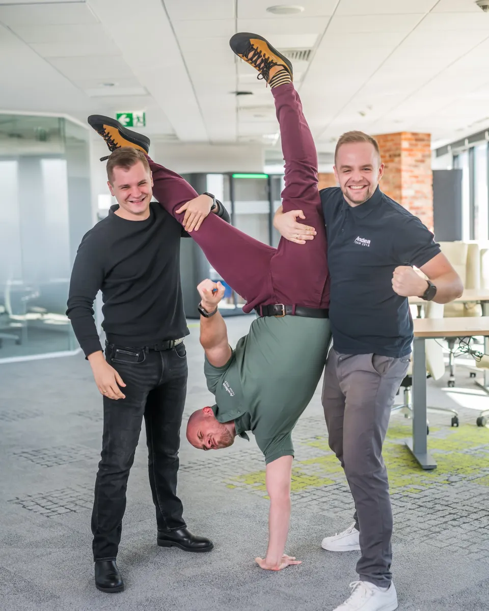 Three smiling men in an office setting. One man is doing a handstand supported by two others, forming a human pyramid. One of them wears a shirt with Andea logo, suggesting team spirit and problem-solving.