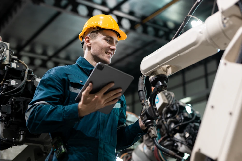 Smiling male factory worker in a blue jumpsuit and yellow hard hat holds a tablet and inspects a robotic arm. Modern manufacturing, production management.