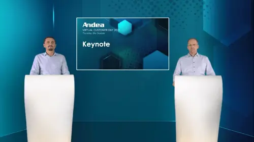 Two men in light blue shirts stand at white podiums in front of a teal background with hexagonal graphics. A screen displays 'Andea Virtual Customer Day 2021 Keynote'. Digital transformation for manufacturing.