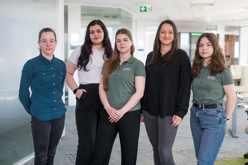 Five diverse women in casual business attire stand together in a bright, modern office. MES and APS software company team.