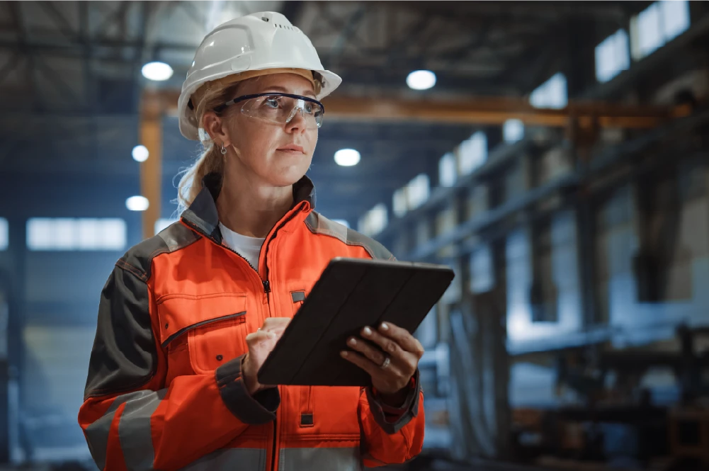 A determined woman wearing a white hard hat and orange safety jacket with reflective stripes holds a tablet computer in a manufacturing facility. She is looking forward with concentration, suggesting production management or data review.