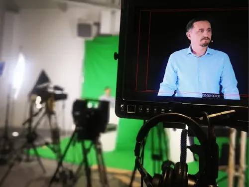 Man in blue shirt on studio monitor screen, production cameras, green screen studio, and headphones in foreground for manufacturing MES APS digital transformation software demo.