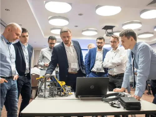 Diverse group of men in business attire gathered around a table, intently observing a laptop displaying software and a complex robotic toy. Focus is on production planning and manufacturing solutions.