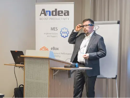 A man in a dark suit and white shirt adjusts smart glasses at a conference podium with laptops and a whiteboard in background. Andea logo visible.