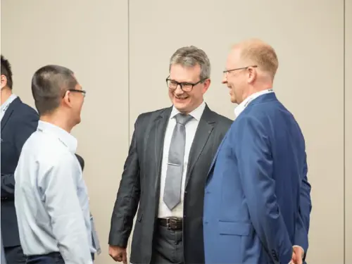 Three men in suits talking at a business event. Two men are smiling while engaged in conversation. This image relates to manufacturing software solutions and digital transformation.