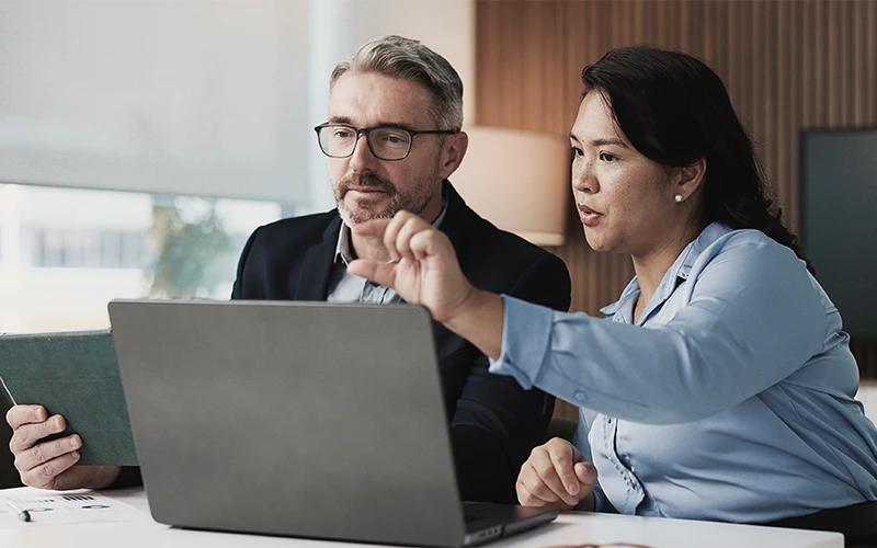 Two manufacturing professionals, a man with glasses and a woman, collaboratively review production data on a laptop. The woman points at the screen, indicating a focus on MES or APS solutions for manufacturing efficiency.