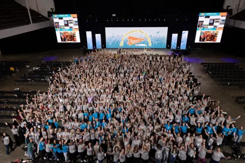 Large audience with hands raised celebrating at Summit 2024. Digital transformation, MES, and APS solutions highlighted on screens. Energetic manufacturing industry event.