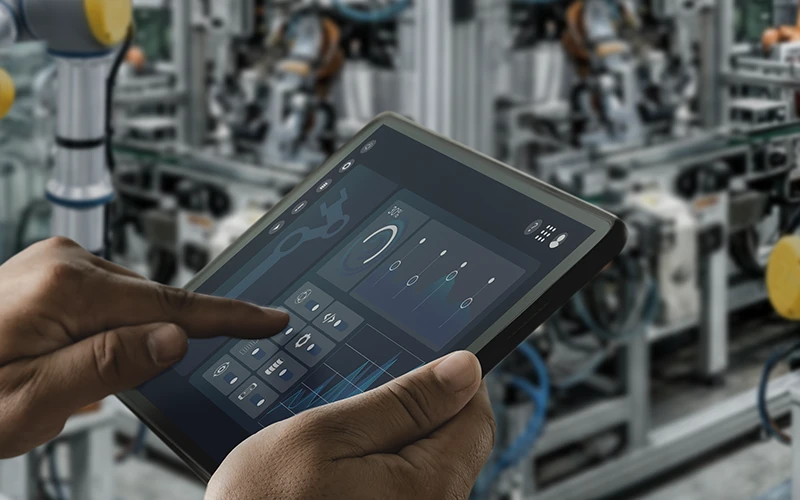 Hands operate a tablet displaying an industrial control interface with graphs and icons. A robotic arm and manufacturing equipment are blurred in the background.